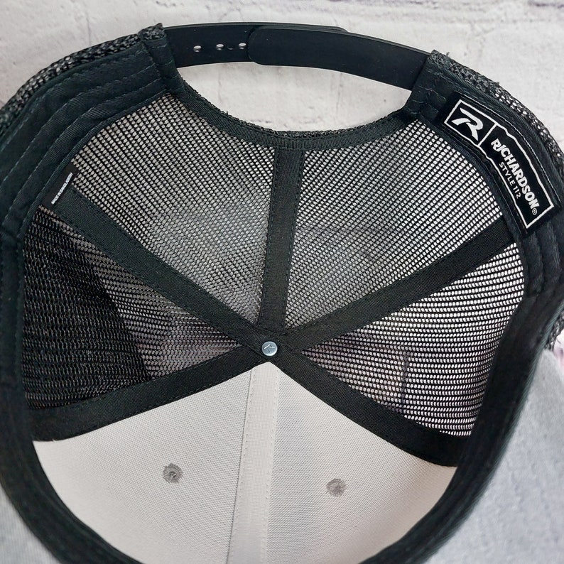 May include: A close-up view of the underside of a black and gray trucker hat. The hat features a black mesh back, a gray front panel, and a black adjustable snap closure. The brand name "RICHARDSON" is visible on a tag.