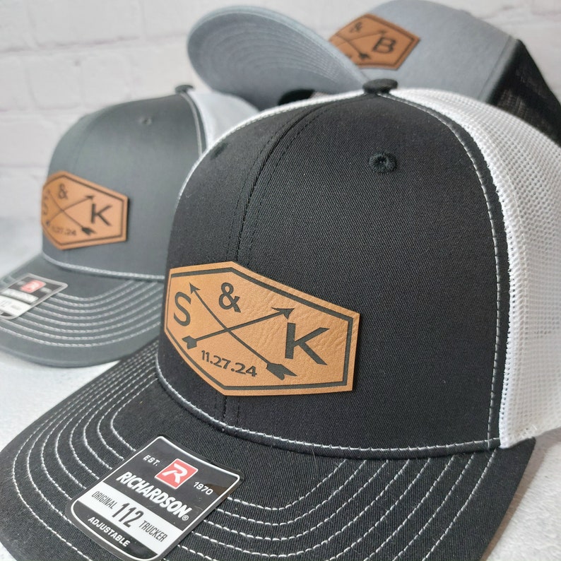 May include: Black and white trucker hat with a brown leather patch that says "S&K 11.27.24". The patch has two arrows crossing each other. The hat has a Richardson logo and says "Original 112 Trucker Adjustable".