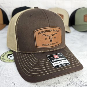 May include: Brown trucker hat with a tan mesh back and a brown brim. The hat features a leather patch with the text "LONGHORN RANCH BLACK ROCK, TX" and a steer skull graphic. The hat is adjustable and has a Richardson tag.