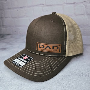 Op de afbeelding: Bruine en beige trucker cap met een bruin leren patch waarop "DAD ESTABLISHED 2025" staat.