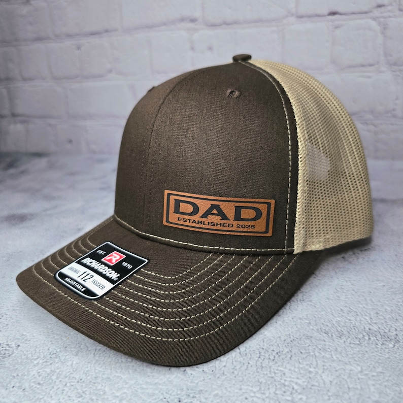 Op de afbeelding: Bruine en beige trucker cap met een bruin leren patch waarop "DAD ESTABLISHED 2025" staat.