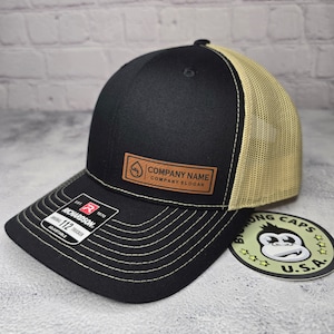 May include: Black and tan trucker hat with a curved brim and mesh back. Features a brown leather patch with the text "COMPANY NAME COMPANY SLOGAN". The hat has a Richardson 112 Trucker tag.
