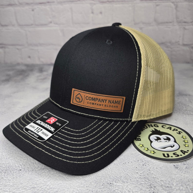 May include: Black and tan trucker hat with a curved brim and mesh back. Features a brown leather patch with the text "COMPANY NAME COMPANY SLOGAN". The hat has a Richardson 112 Trucker tag.
