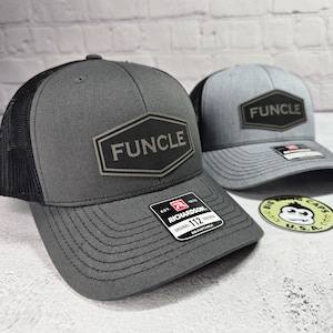 May include: Two gray trucker hats with black mesh back panels. Both hats feature a black hexagonal patch with the word "FUNCLE" in white lettering. The hat on the left has a black and red adjustable strap with the words "Richardson 112" printed on it. The hat on the right has a gray adjustable strap.