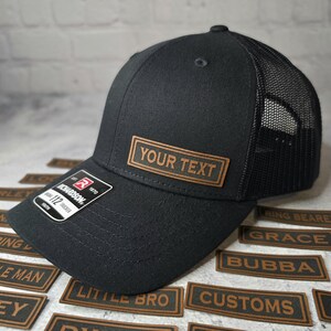 以下が含まれることがあります： 黒と茶色のトラッカーハット。茶色の革のパッチに「YOUR TEXT」と書いてあります。