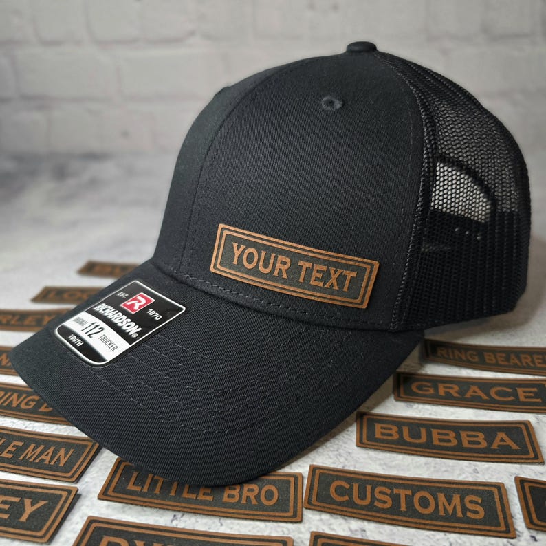 以下が含まれることがあります： 黒と茶色のトラッカーハット。茶色の革のパッチに「YOUR TEXT」と書いてあります。