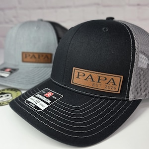 May include: Black trucker hat with a gray mesh back and a brown leather patch that reads "PAPA EST. 2026". The hat has a curved brim and white stitching. A gray hat with a similar design is in the background.