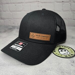 May include: Black trucker hat with a mesh back and a curved brim. The hat features a leather patch with the text "YOUR COMPANY YOUR CUSTOM TEXT". A Richardson 112 adjustable tag is on the brim.