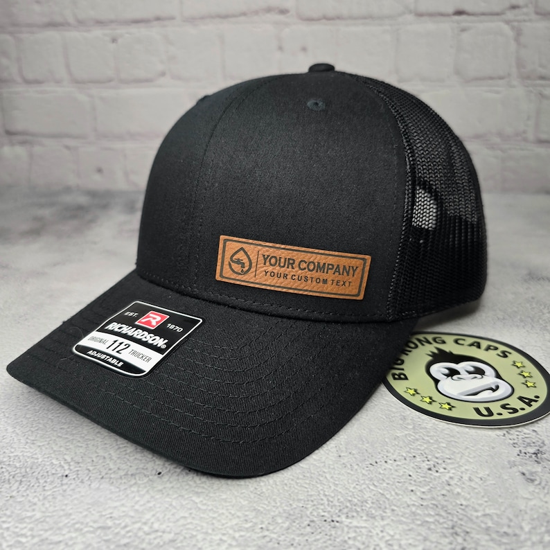 May include: Black trucker hat with a mesh back and a curved brim. The hat features a leather patch with the text "YOUR COMPANY YOUR CUSTOM TEXT". A Richardson 112 adjustable tag is on the brim.