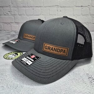 May include: Two gray trucker hats with black mesh back panels. The hats have a brown leather patch with the word "GRANDPA" printed in white.