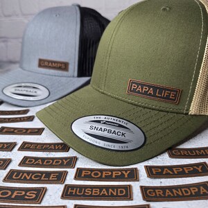 May include: Two baseball caps with different colored fabric and mesh panels. The caps have a brown leather patch with the text "GRAMPS" and "PAPA LIFE" on them. The caps are surrounded by small brown leather patches with different words on them, such as "POPPY", "HUSBAND", and "GRANDPA".