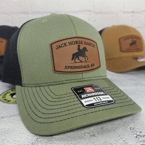 May include: An olive green trucker hat with a black mesh back and a brown leather patch. The patch features the text "JACK HORSE RANCH SPRINGDALE, AR" and a silhouette of a rider on a horse. The hat has a curved brim.