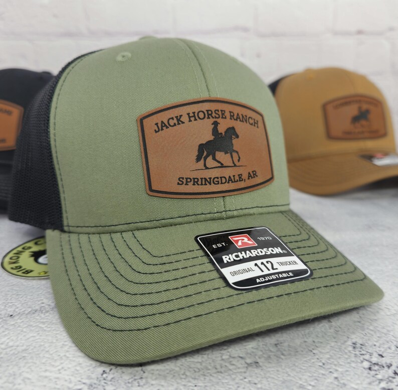 May include: An olive green trucker hat with a black mesh back and a brown leather patch. The patch features the text "JACK HORSE RANCH SPRINGDALE, AR" and a silhouette of a rider on a horse. The hat has a curved brim.