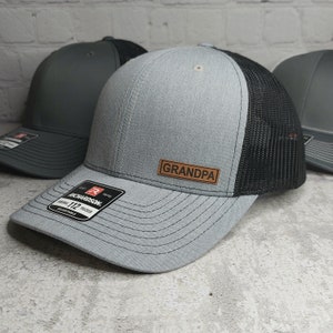May include: A gray trucker hat with a black mesh back and a brown leather patch that reads "GRANDPA". The hat is a Richardson 112 model, adjustable, and has a curved brim.
