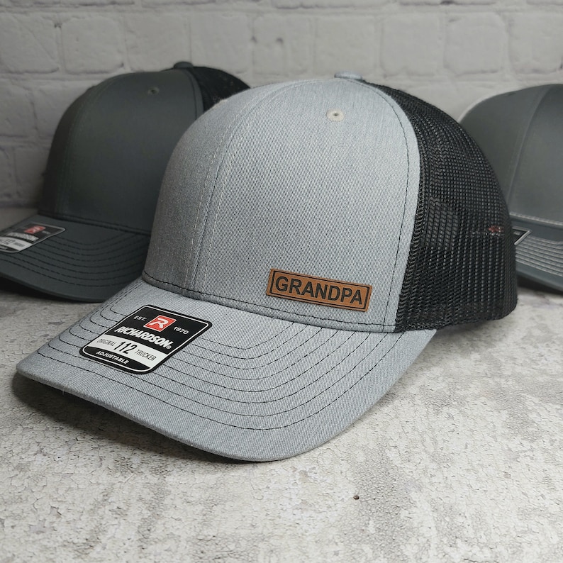 May include: A gray trucker hat with a black mesh back and a brown leather patch that reads "GRANDPA". The hat is a Richardson 112 model, adjustable, and has a curved brim.