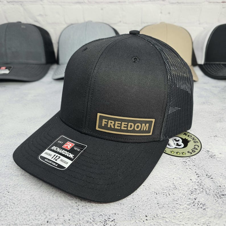 May include: Black trucker hat with a mesh back and a curved brim. The hat features a rectangular patch with the word "FREEDOM" in gold lettering. The hat is adjustable and has a Richardson tag.