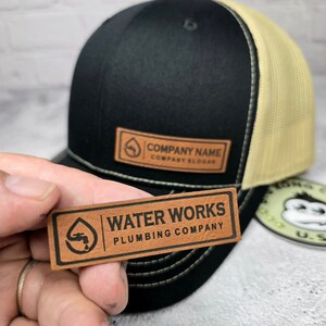 May include: Black and tan trucker hat with a leather patch. The patch reads "COMPANY NAME" and "COMPANY SLOGAN". A second leather patch with the text "WATER WORKS PLUMBING COMPANY" is held in front of the hat.