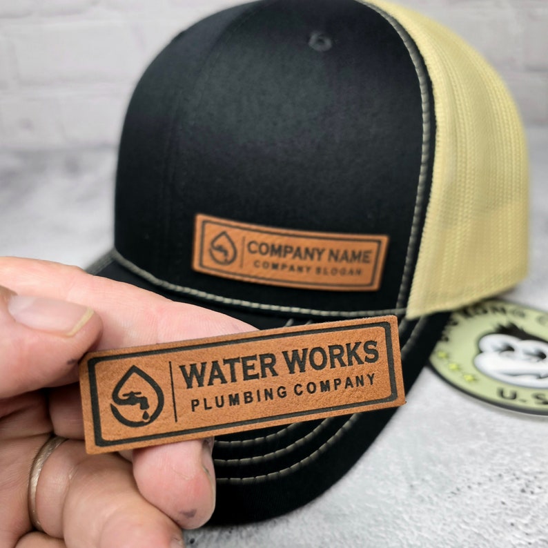 May include: Black and tan trucker hat with a leather patch. The patch reads "COMPANY NAME" and "COMPANY SLOGAN". A second leather patch with the text "WATER WORKS PLUMBING COMPANY" is held in front of the hat.