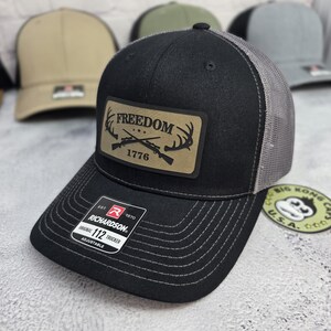 May include: Black trucker hat with a gray mesh back and a curved brim. The hat features a rectangular patch with the word "FREEDOM" above crossed rifles and antlers, with "1776" below. The hat is adjustable and has the "RICHARDSON" brand label.