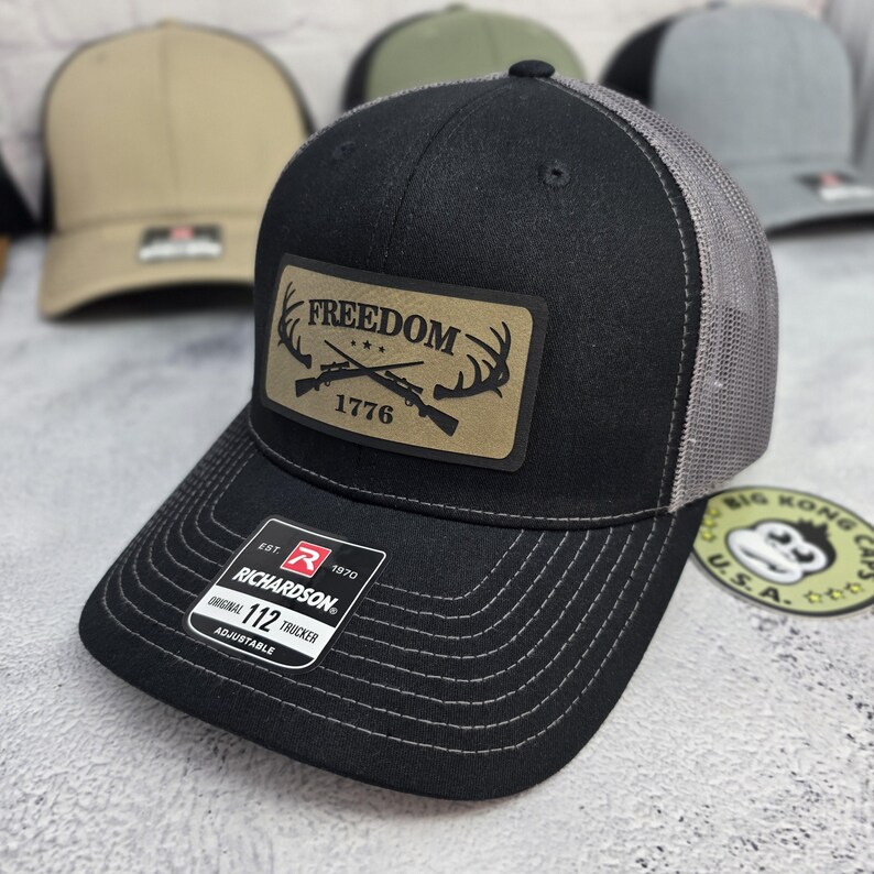 May include: Black trucker hat with a gray mesh back and a curved brim. The hat features a rectangular patch with the word "FREEDOM" above crossed rifles and antlers, with "1776" below. The hat is adjustable and has the "RICHARDSON" brand label.