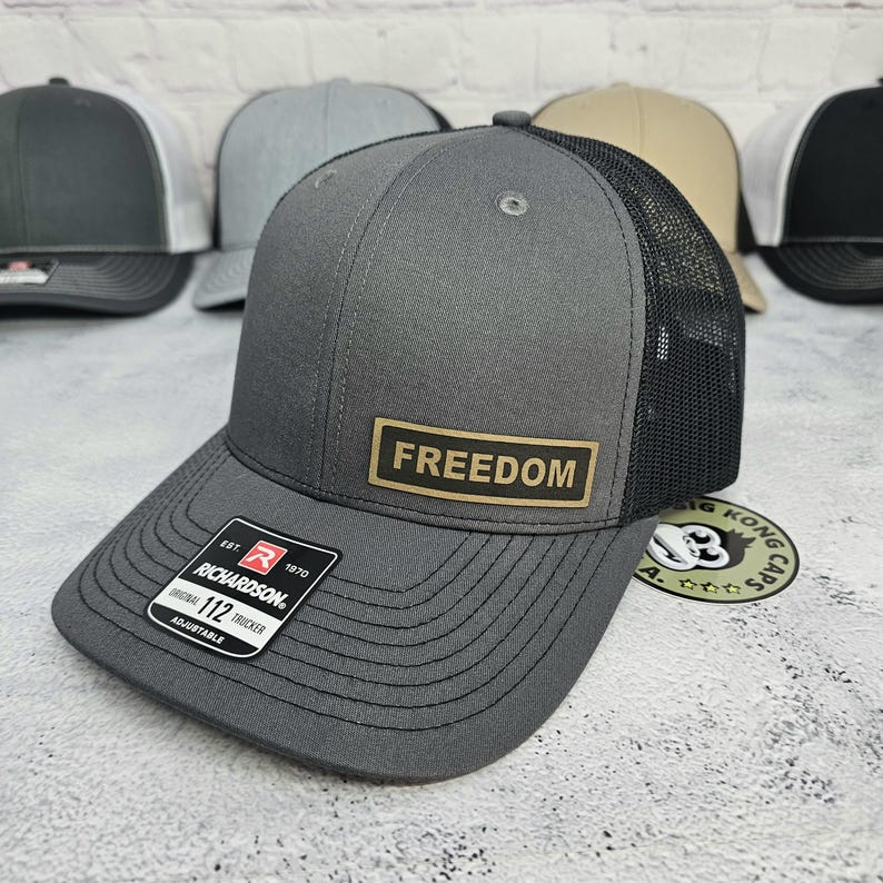 May include: A gray trucker hat with a black mesh back and a curved brim. The hat features a rectangular patch with the word "FREEDOM" in gold lettering. The hat is adjustable and has a Richardson 112 tag.