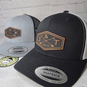 May include: Two black and gray trucker hats with leather patches. The patch on the left hat has the letters "B & K" with a date. The patch on the right hat has the letters "J & T" with a date. Both hats have a YUPOONG SNAPBACK label.