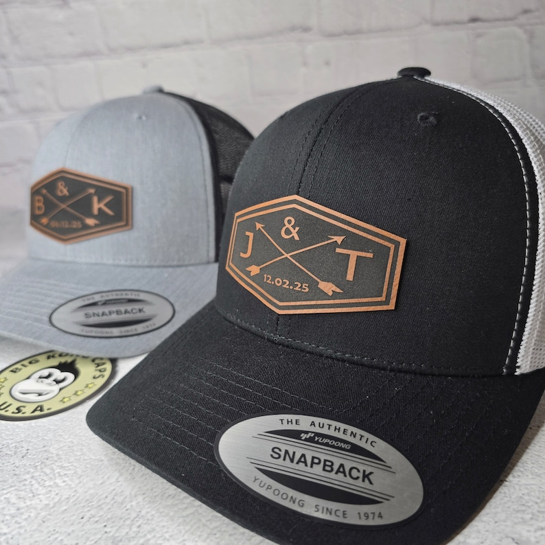 May include: Two black and gray trucker hats with leather patches. The patch on the left hat has the letters "B & K" with a date. The patch on the right hat has the letters "J & T" with a date. Both hats have a YUPOONG SNAPBACK label.