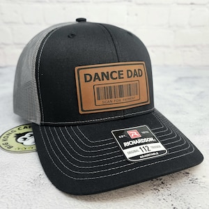 May include: Black trucker hat with a gray mesh back and a curved brim. The hat features a brown leather patch with the words "DANCE DAD" and a barcode. The hat is adjustable.