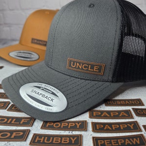 May include: A gray and black trucker hat with a brown leather patch that says "UNCLE". The hat is surrounded by brown leather patches with various words, including "PAPPY", "POPPY", "HUBBY", and "PEEPAW".