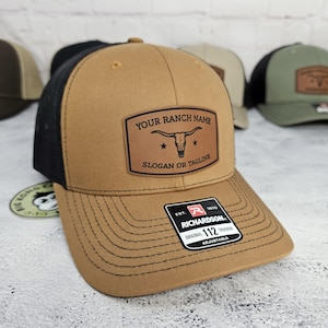 May include: A brown trucker hat with a black mesh back. The hat features a leather patch with the text "YOUR RANCH NAME" above a steer skull graphic and "SLOGAN OR TAGLINE" below. The hat is adjustable and has a Richardson tag.
