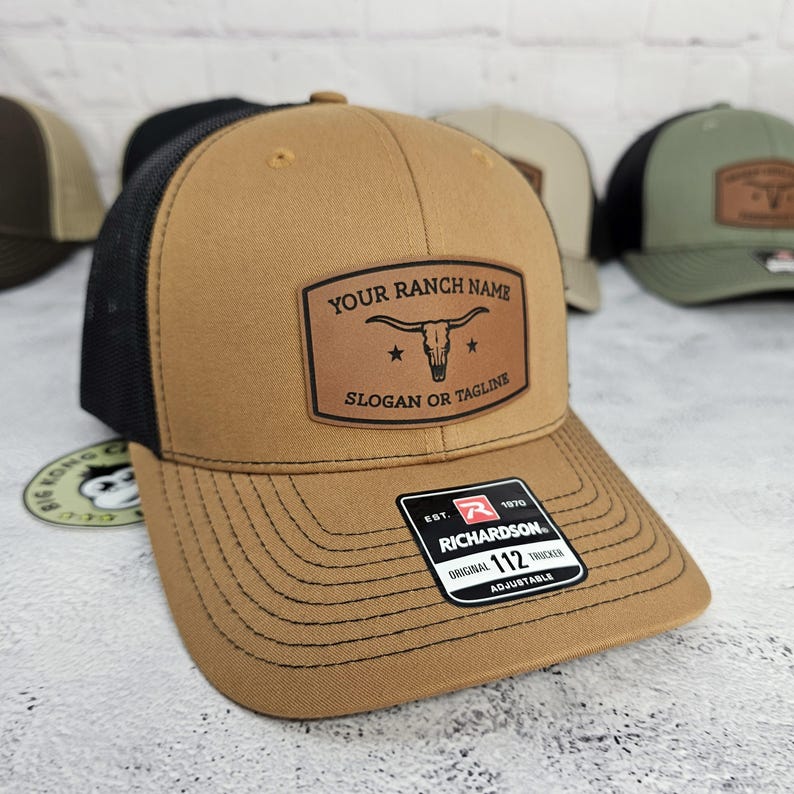 May include: A brown trucker hat with a black mesh back. The hat features a leather patch with the text "YOUR RANCH NAME" above a steer skull graphic and "SLOGAN OR TAGLINE" below. The hat is adjustable and has a Richardson tag.