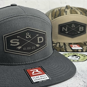May include: Two flat bill snapback hats. One is gray with a black mesh back and a custom logo with the letters "S" and "D" and the date "10.02.25". The other hat has a camouflage pattern and a custom logo with the letters "N" and "B" and the date "12.22.26".