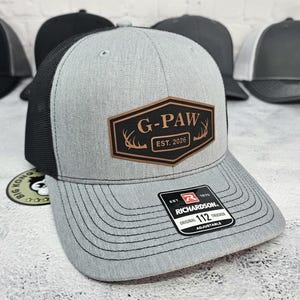 May include: A gray trucker hat with a black mesh back and a curved brim. The hat features a brown patch with the text "G-PAW EST. 2026" and a Richardson patch. The hat is adjustable.