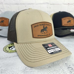 May include: Tan trucker hat with a brown mesh back and a leather patch that reads "Jack Horse Ranch Springdale, AR." The hat has a curved brim and the Richardson 112 Trucker tag.