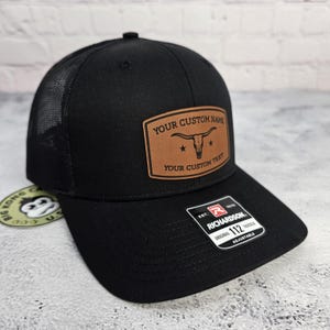 May include: Black trucker hat with a mesh back and a curved brim. A brown leather patch on the front displays a longhorn skull and the text "YOUR CUSTOM NAME" and "YOUR CUSTOM TEXT". The hat is adjustable.