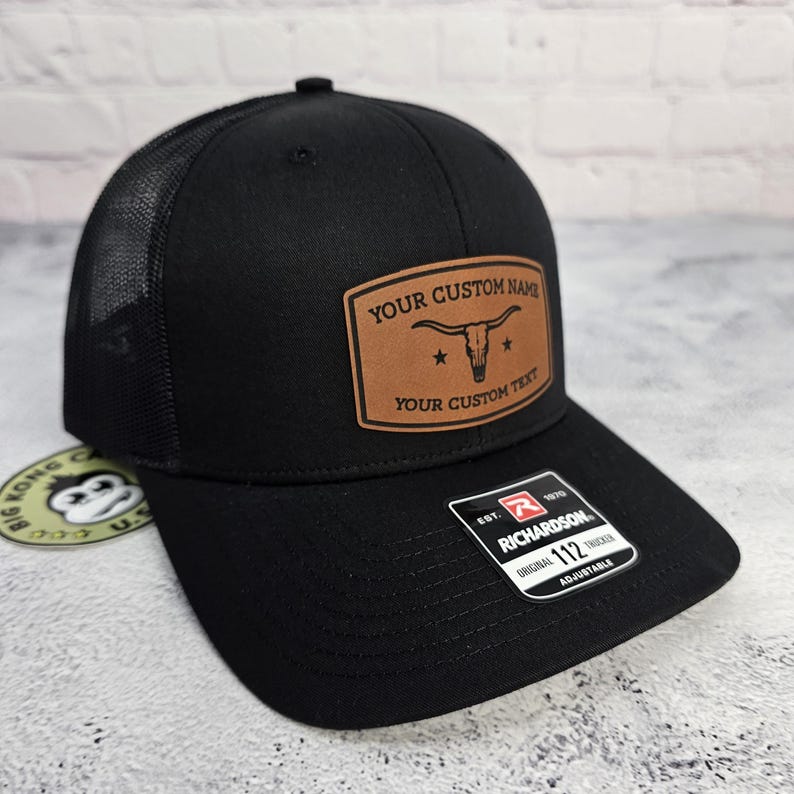 May include: Black trucker hat with a mesh back and a curved brim. A brown leather patch on the front displays a longhorn skull and the text "YOUR CUSTOM NAME" and "YOUR CUSTOM TEXT". The hat is adjustable.