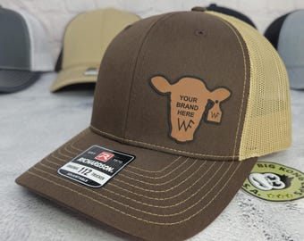 May include: Brown trucker hat with a tan mesh back and a curved brim. The hat features a leather patch with a cow graphic and the text "YOUR BRAND HERE". The hat is adjustable and has the Richardson brand label.