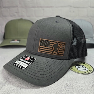 May include: A gray and black trucker hat with a brown leather patch featuring an American flag and a silhouette of Bigfoot holding an arrow. The hat is adjustable and has the Richardson brand logo on the side.