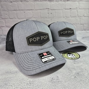 May include: Two gray and black trucker hats with a black patch that says "POP POP" on each. The hats are on a brick background.