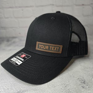 以下が含まれることがあります： 「YOUR TEXT」と書かれた茶色の革のパッチが付いた黒いトラッカーハットです。