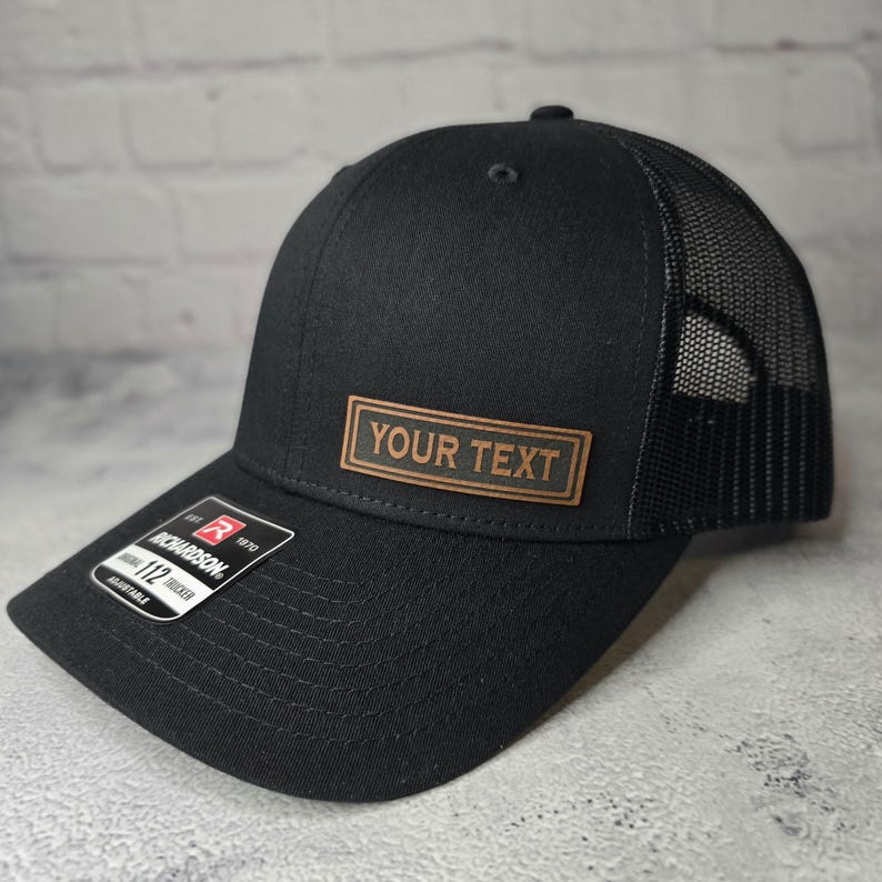 以下が含まれることがあります： 「YOUR TEXT」と書かれた茶色の革のパッチが付いた黒いトラッカーハットです。