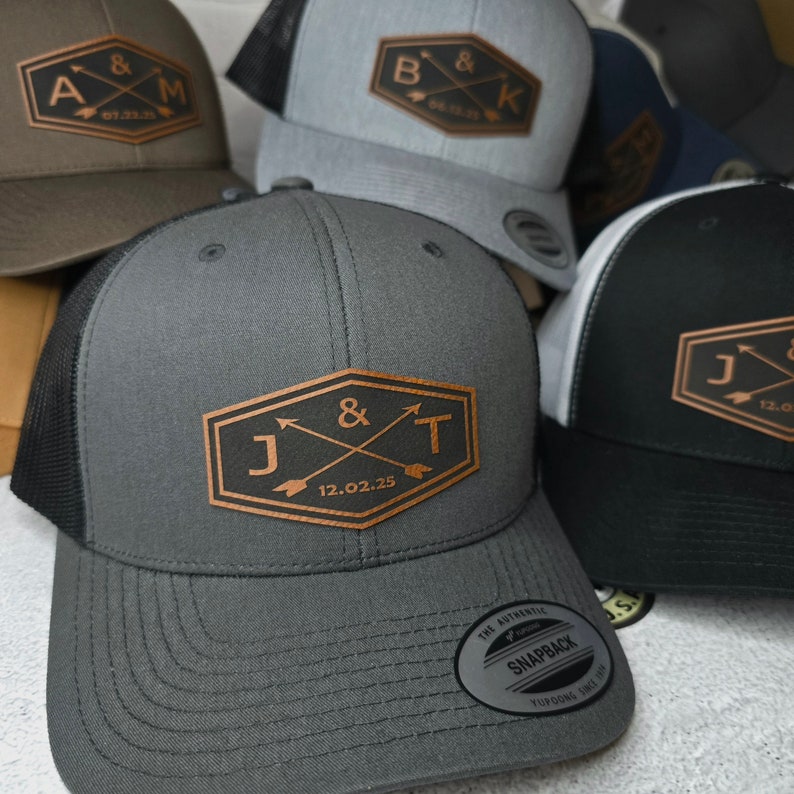 May include: A close-up of a gray trucker hat with a brown leather patch that says "J & T 12.02.25" with two arrows crossing each other. The hat has a black mesh back and a black brim.