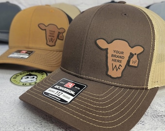 May include: Brown and tan trucker hats with a cow graphic and the text "YOUR BRAND HERE". The hats have a Richardson 112 Trucker label and are adjustable. The hats are on a white surface.
