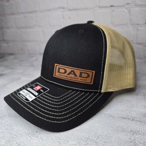 Op de afbeelding: Zwarte en beige truckercap met een bruin leren patch waarop "DAD ESTABLISHED 2025" staat.