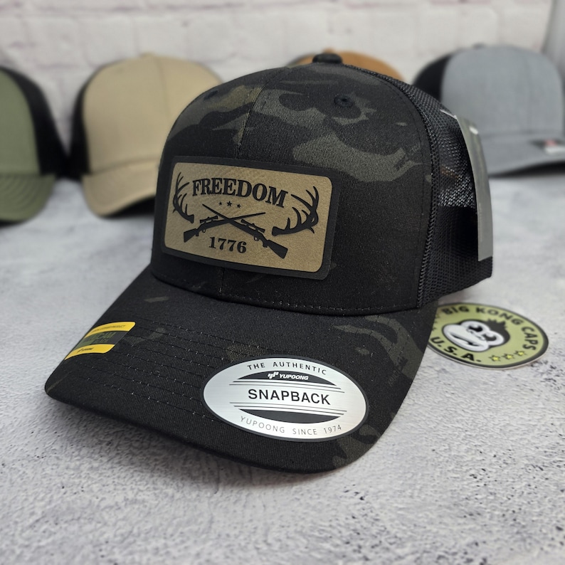 May include: Black camouflage baseball cap with a mesh back. The cap features a leather patch with the word "FREEDOM", crossed rifles, and the year "1776". The cap also has a curved brim and a snapback closure.