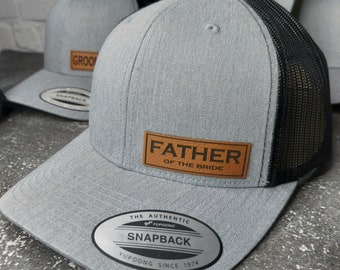 May include: A gray and black mesh baseball cap with a brown leather patch that says "Father of the Bride". The cap has a black mesh back and a gray front with a white sticker that says "The Authentic Yupoong Snapback Yupoong Since 1974".