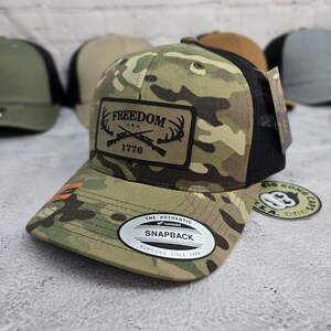 May include: Camouflage pattern snapback hat with a black mesh back. The front features a patch with the word "FREEDOM", crossed rifles, and the year "1776". The hat has a curved brim and a "Yupoong" tag.
