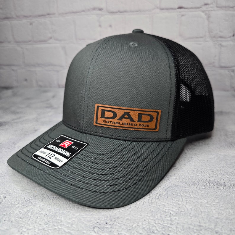 Op de afbeelding: Een grijze en zwarte trucker-pet met een bruin leren patch waarop "DAD ESTABLISHED 2025" staat.