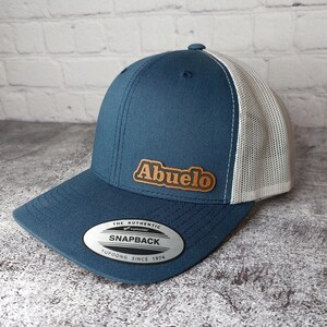 May include: A blue and white trucker hat with the word "Abuelo" in a brown leather patch. The hat has a curved brim and a snapback closure. The tag reads "Snapback" and "Yupoong Since 1974".