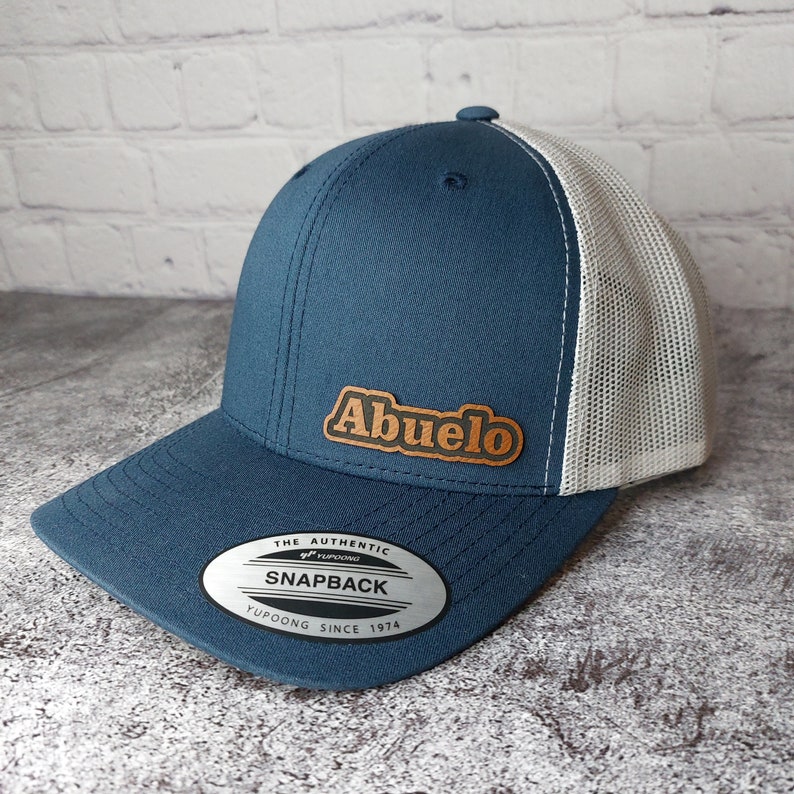 May include: A blue and white trucker hat with the word "Abuelo" in a brown leather patch. The hat has a curved brim and a snapback closure. The tag reads "Snapback" and "Yupoong Since 1974".